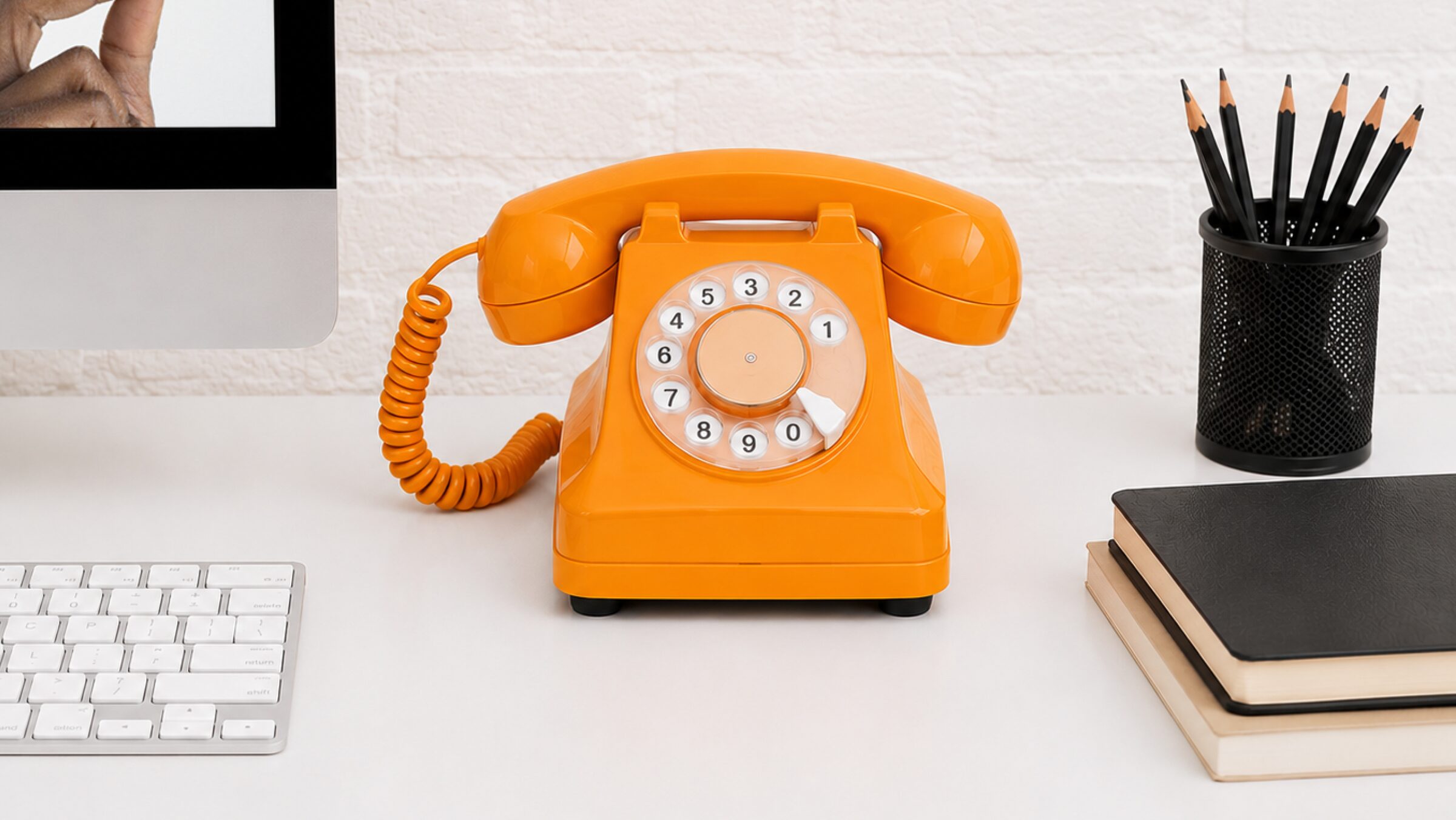 A vintage orange rotary telephone sitting on a desk between a computer monitor, keyboard, notebooks and a pot of pencils